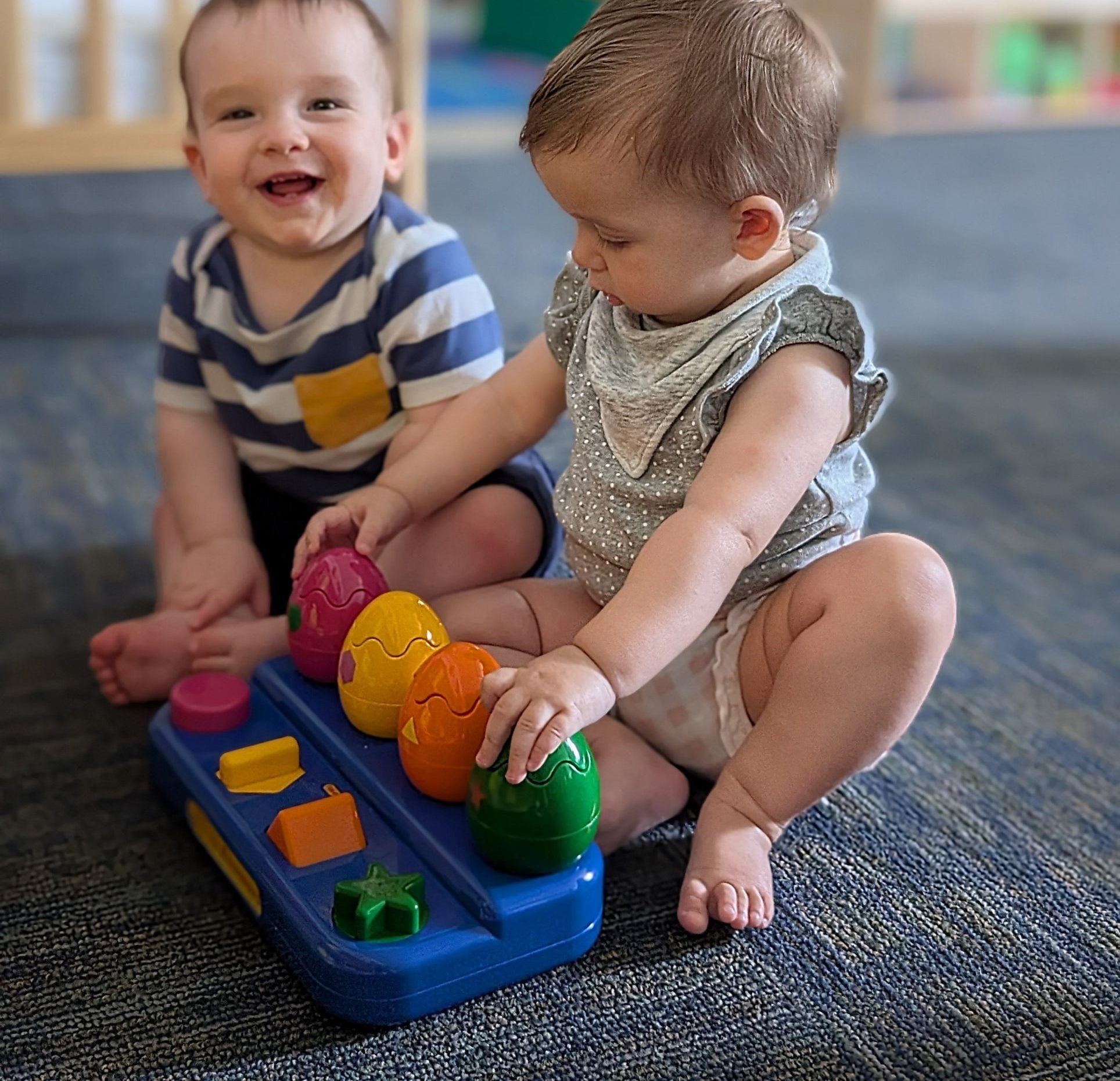Two smiling infants working together to solve an age-appropriate shape toy,